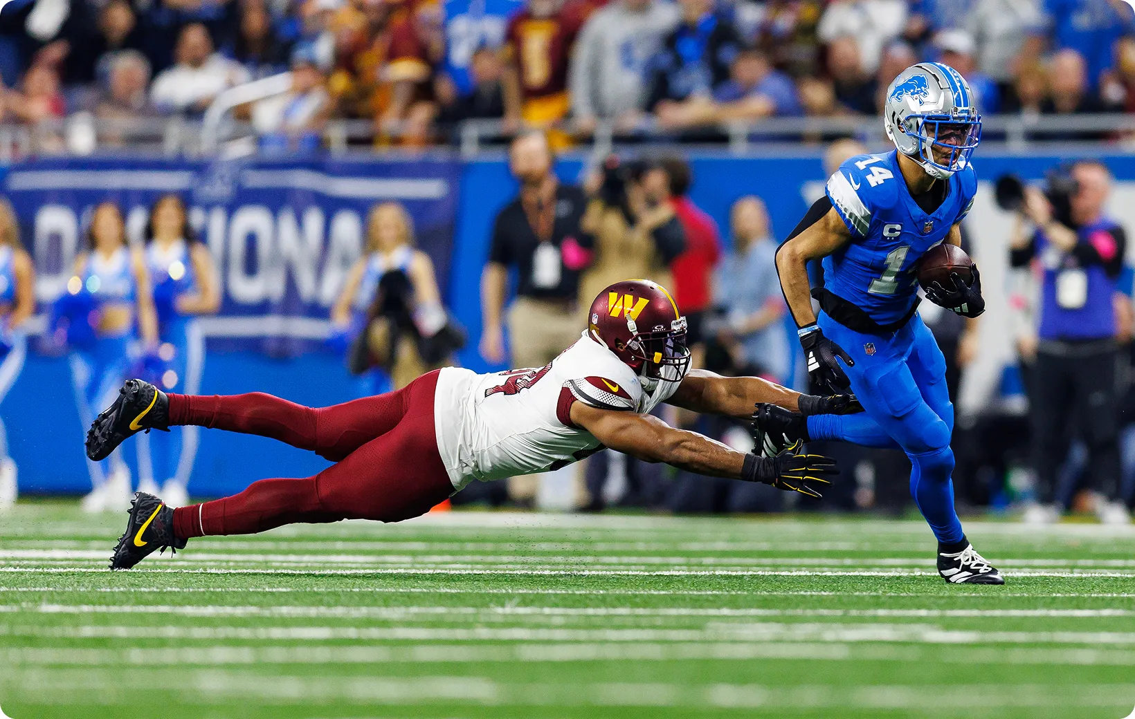 Action-Foto aus einem NFL-Spiel: Ein Footballspieler in blauer Uniform läuft mit dem Ball, während ein Gegenspieler im Hechtsprung versucht, ihn an den Beinen zu tackeln.